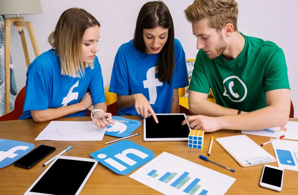 Tres jóvenes analizan datos en una tablet, rodeados de gráficos y dispositivos, mientras visten camisetas de redes sociales.