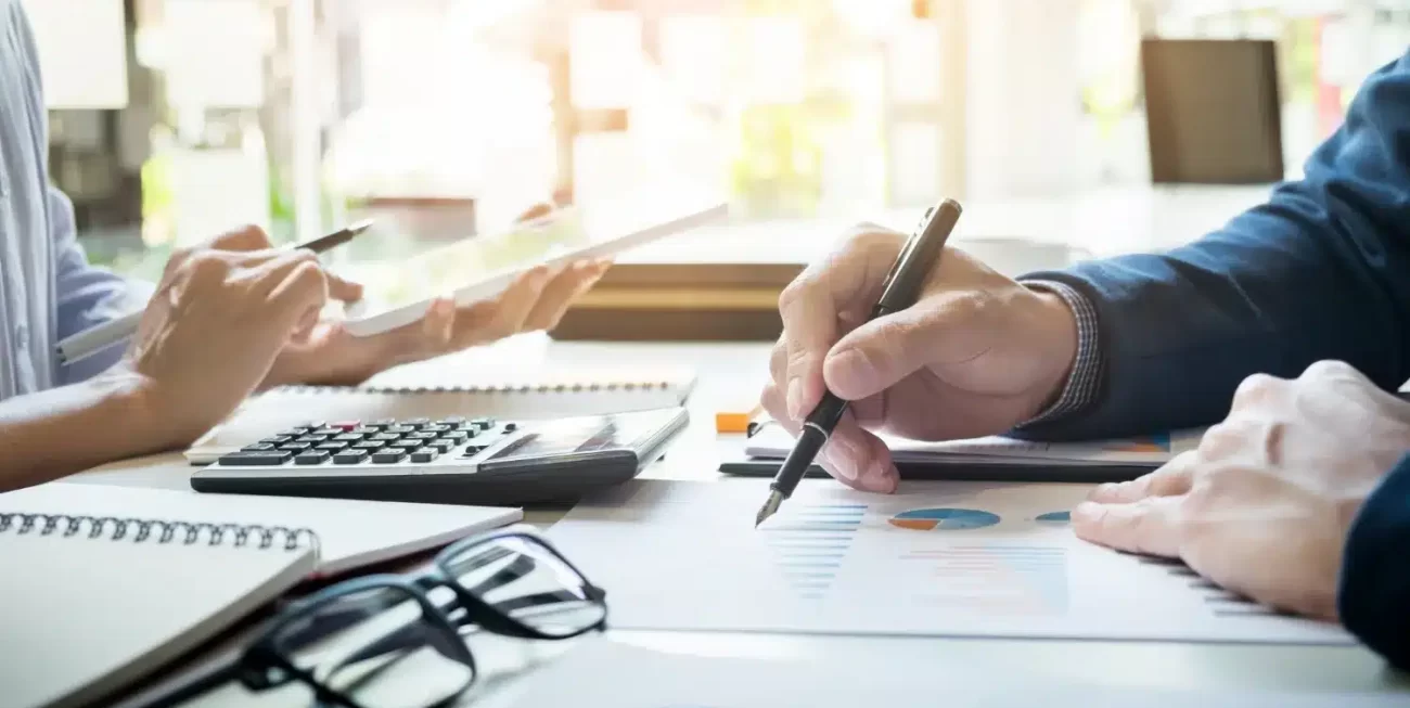 Dos personas colaboran en un análisis de datos, utilizando una tablet, calculadora y documentos con gráficos.