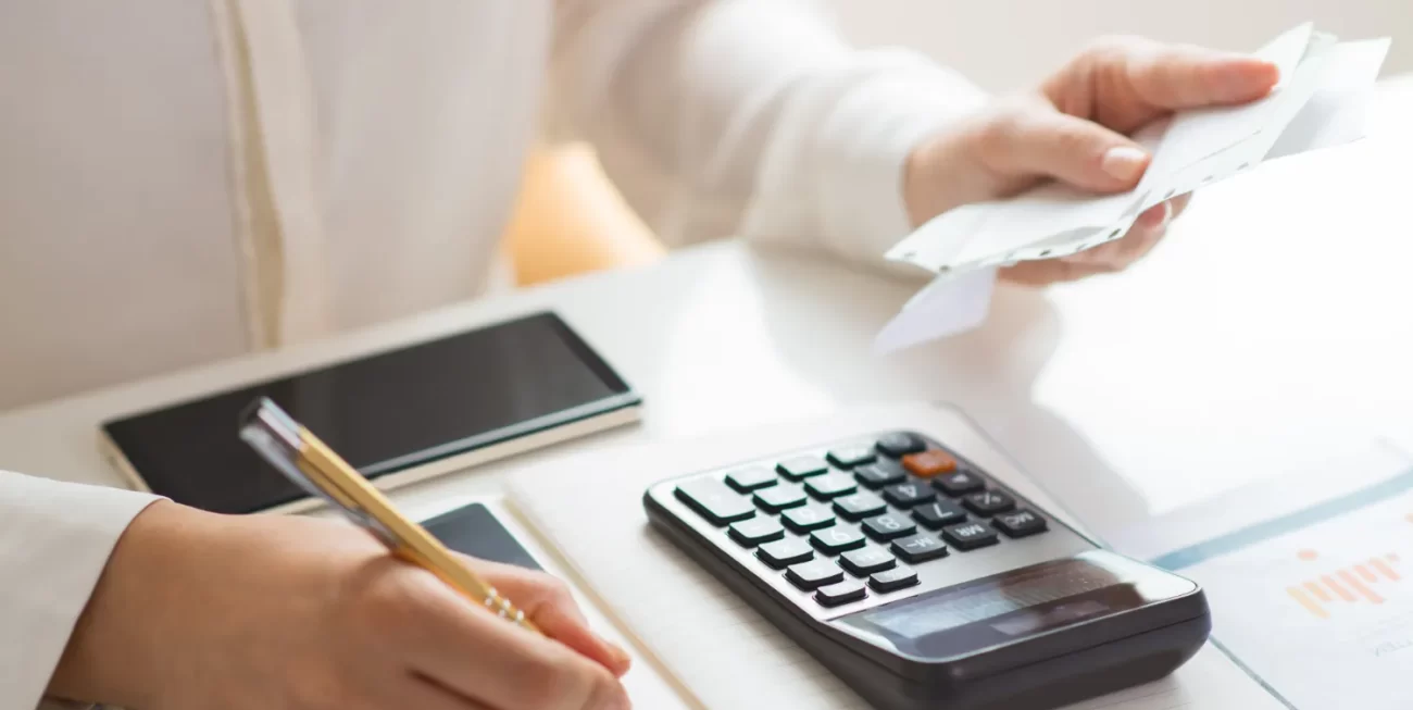 Persona usando una calculadora y tomando notas sobre finanzas en un escritorio con un teléfono móvil al fondo.