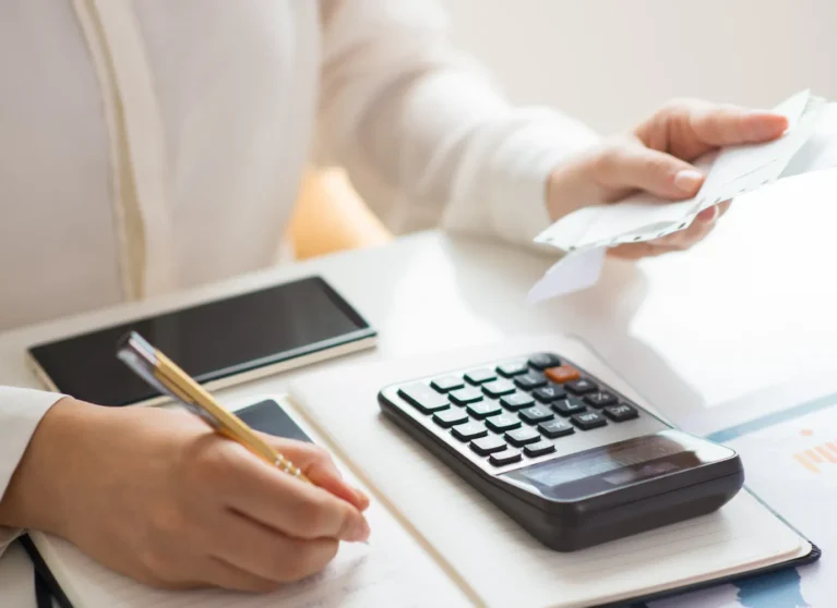 Persona usando una calculadora y tomando notas sobre finanzas en un escritorio con un teléfono móvil al fondo.
