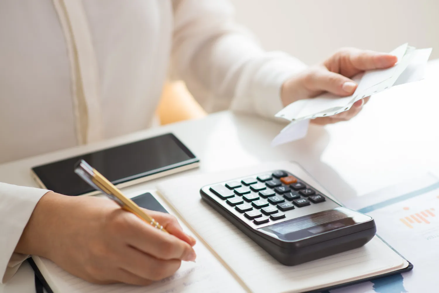 Persona usando una calculadora y tomando notas sobre finanzas en un escritorio con un teléfono móvil al fondo.