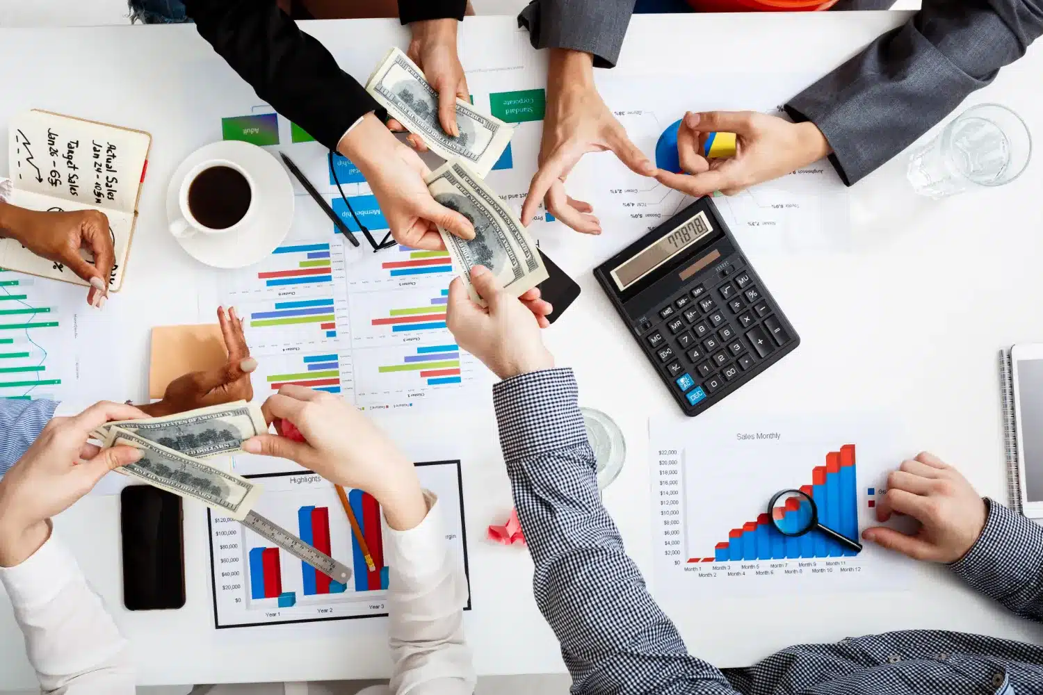 Manos intercambiando dinero sobre una mesa llena de gráficos, calculadora y una taza de café en un entorno de trabajo colaborativo.