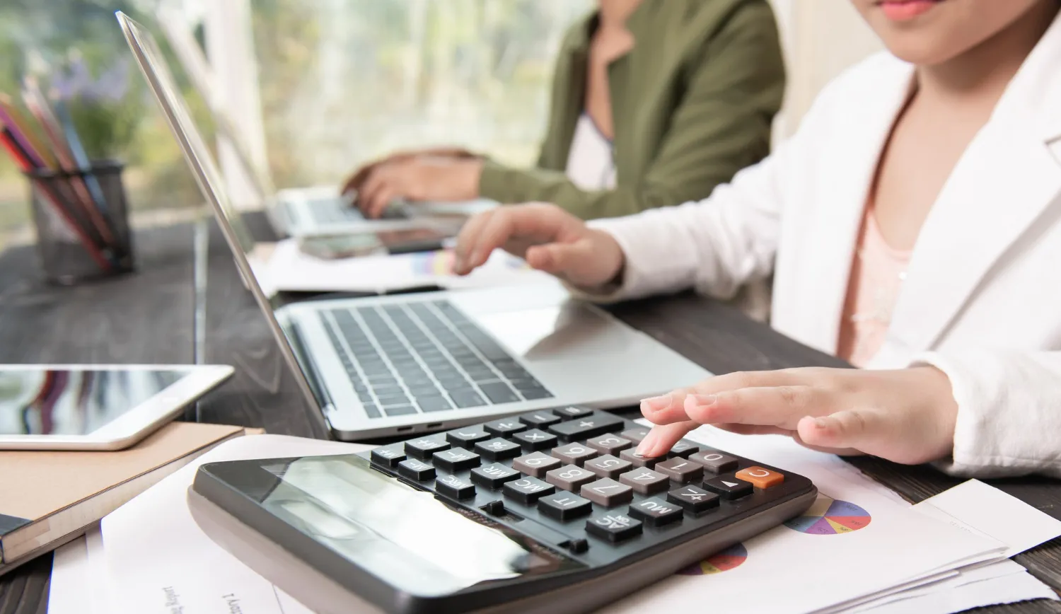 Estudiantes trabajando en tareas financieras con una calculadora y computadoras portátiles en un escritorio.