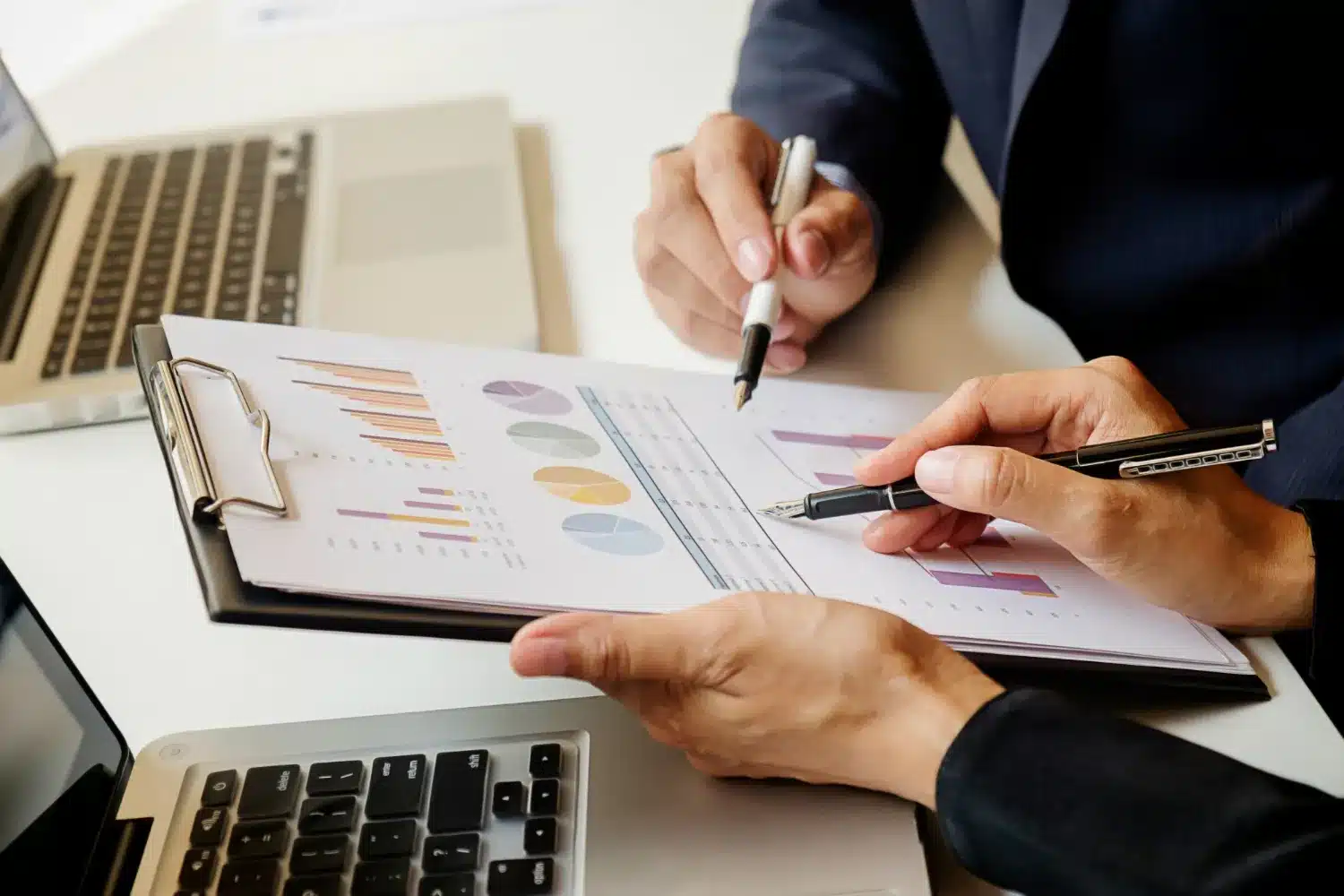 Dos personas analizando gráficos y datos en un documento mientras utilizan computadoras portátiles en una mesa.