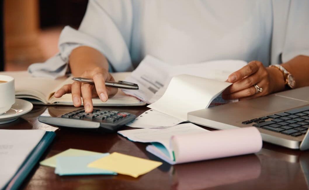 Mujer utilizando una calculadora y revisando documentos financieros en un escritorio con café y computadora portátil.