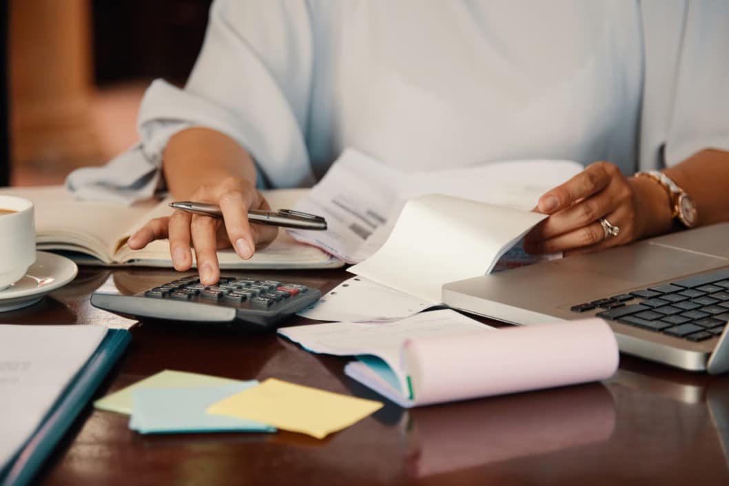 Mujer utilizando una calculadora y revisando documentos financieros en un escritorio con café y computadora portátil.