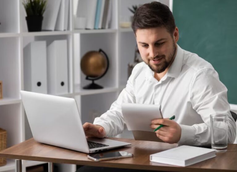 Hombre sonriente revisando documentos mientras trabaja en una computadora portátil en un entorno de oficina organizado.