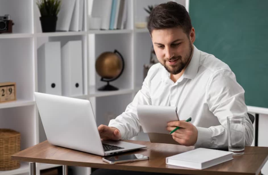 Hombre sonriente revisando documentos mientras trabaja en una computadora portátil en un entorno de oficina organizado.