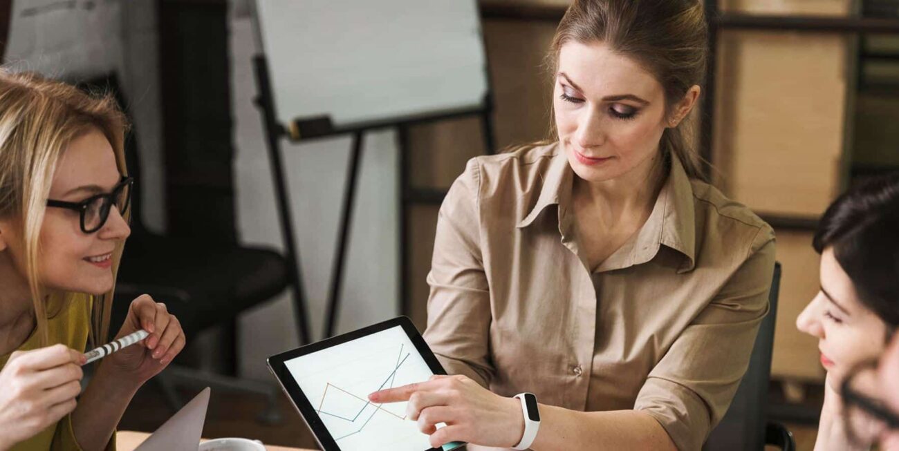 Cuatro mujeres en una reunión analizan datos en una tablet, mostrando gráficos y discutiendo estrategias.